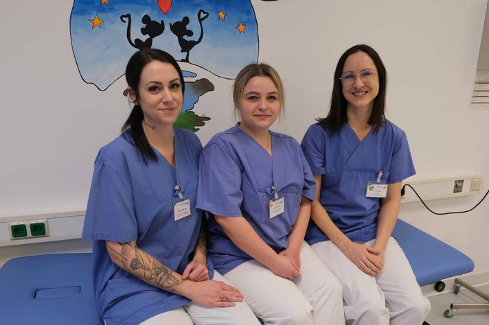 Ein Gruppenfoto mit drei Mitarbeiterinnen der Kindertagesklinik. Alle drei sitzen auf einem Klinikbett und lächeln in die Kamera