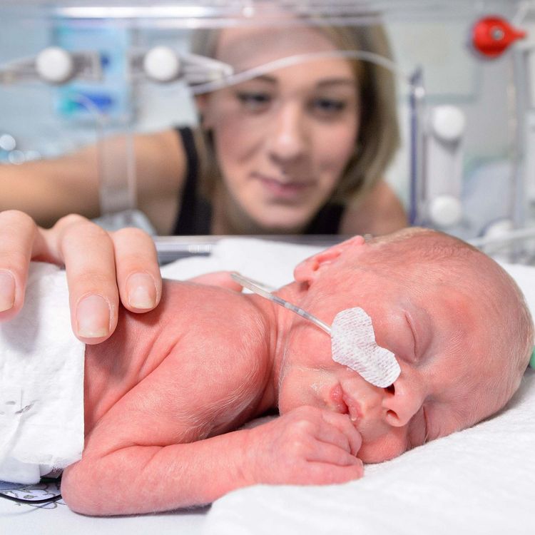 Schlafendes Frühgeborenes im Inkubator, im Hintergrund die Mutter. 