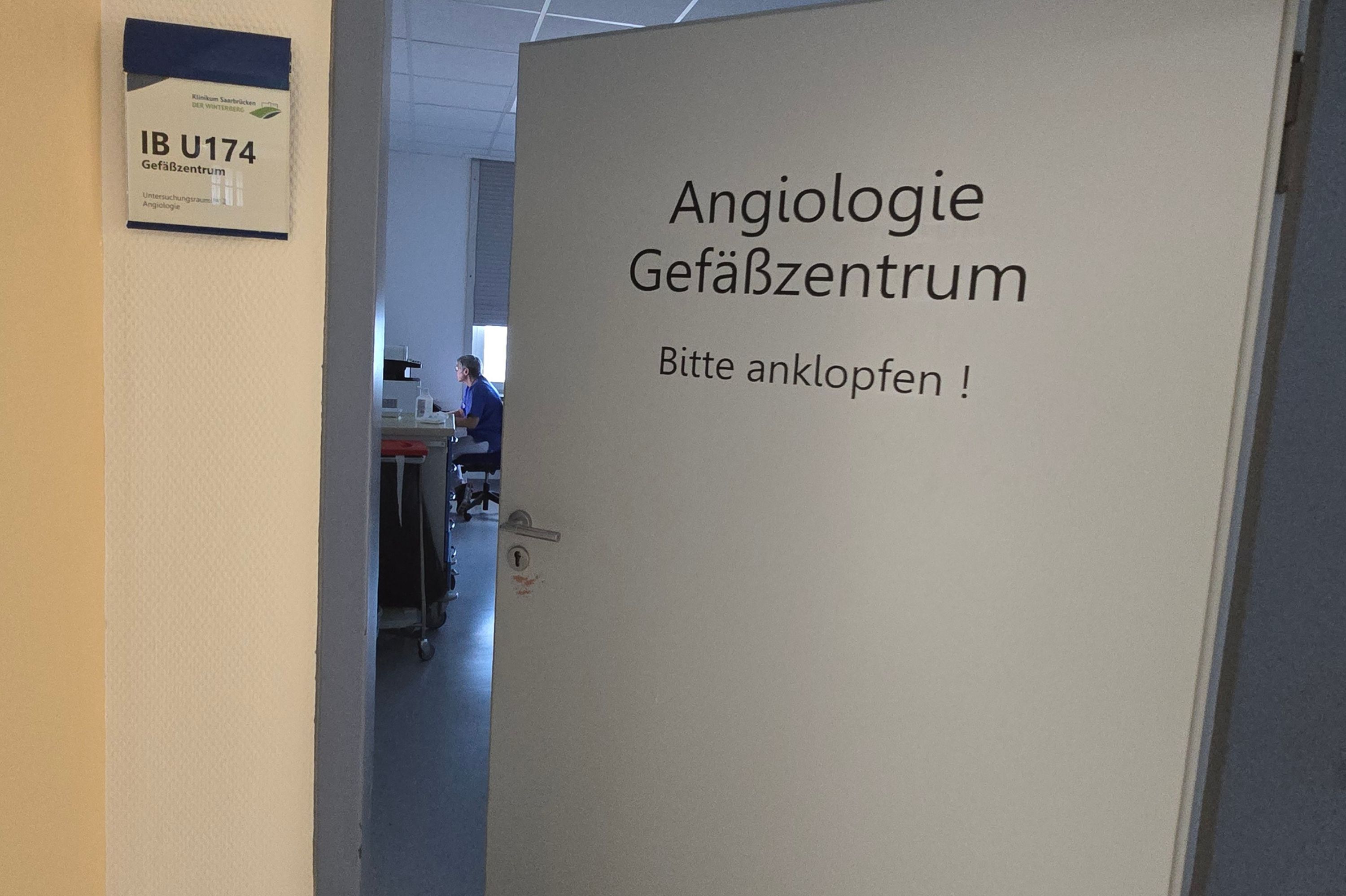 Man sieht eine Tür, die halb geöffnet ist. Im Raum weiter hinten sitzt jemand am Schreibtisch. An der Tür steht "Angiologie".