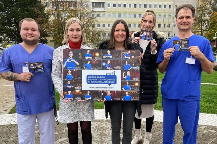 Fünf Vertreter der Recruiting-Kampagne "Die Rolle Deines Lebens" stehen auf dem Hubschrauberlandeplatz vorm Haupteingang des Klinikums Saarbrücken und zeigen eine Collage mit allen Motiven. 