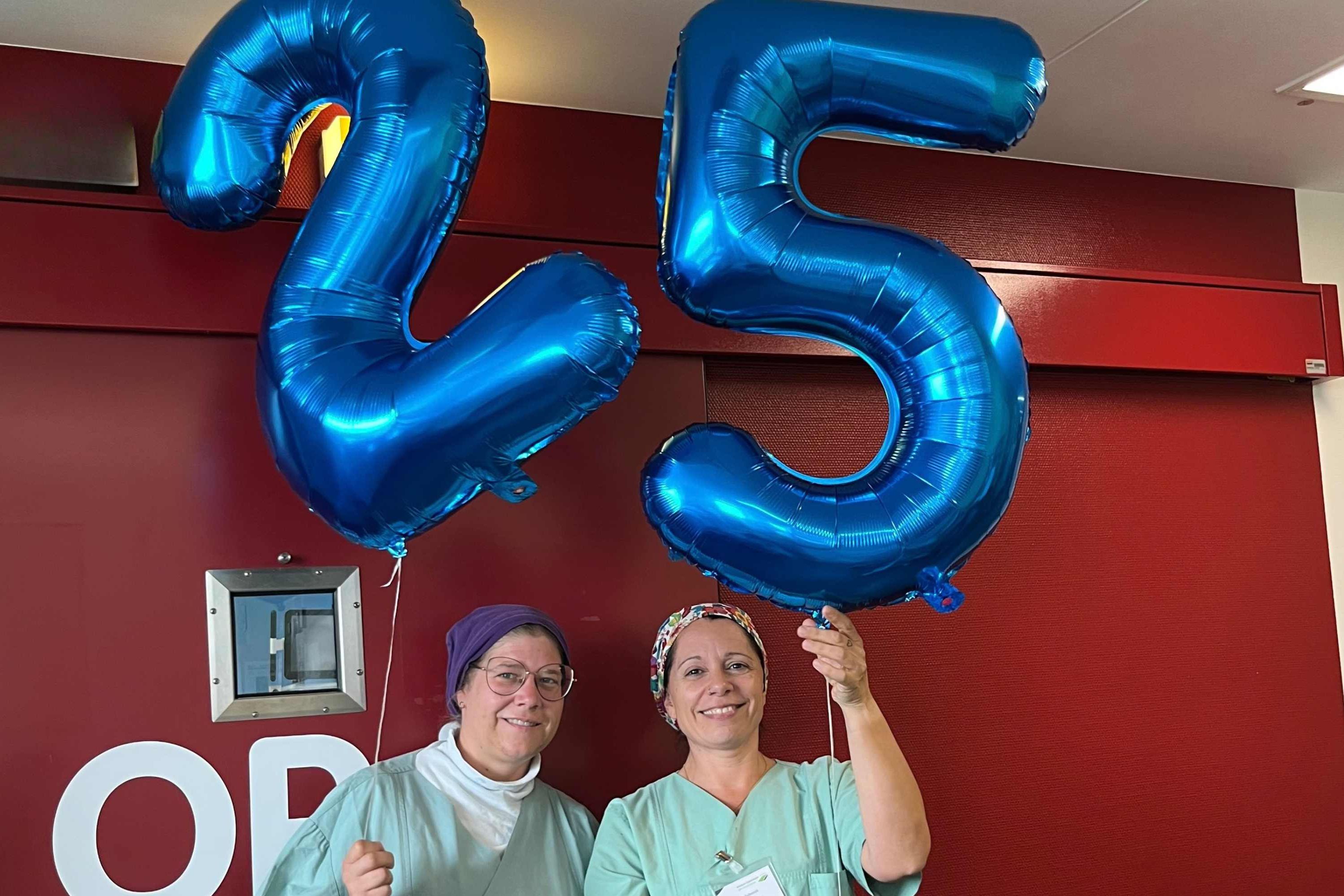 Stefanie Jäckel und Katja Schmitt vor der OP-Tür stehend im Zentral-Op des Klinikums Saarbrücken mit je einem Heliumballon, einer zwei und einer fünf, anlässlich des 25-jährigen Jubiläum.