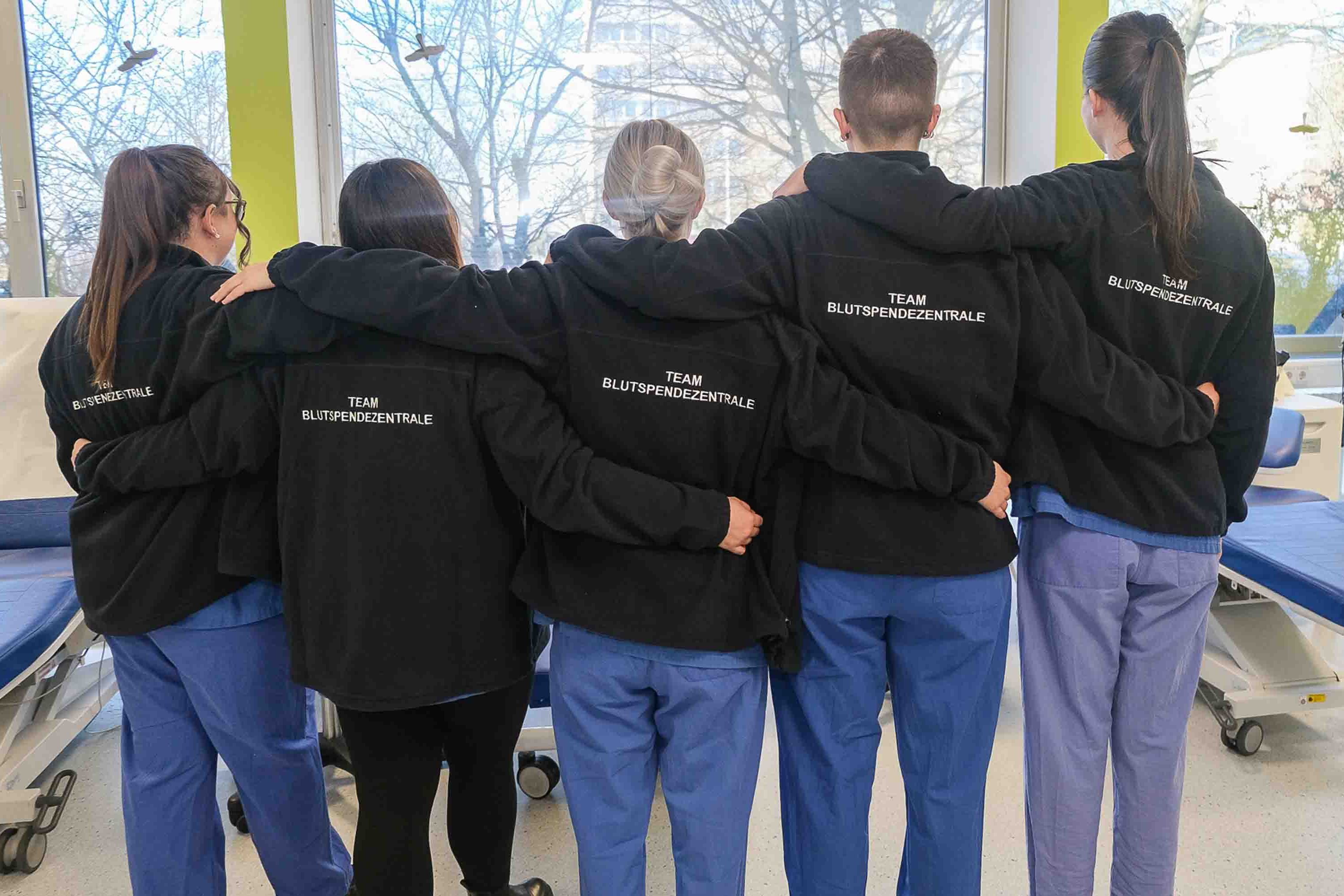 Fünf Kolleginnen der Blutspendezentrale Saar-Pfalz in Saarbrücken stehen mit dem Rücken zur Kamera Arm in Arm da. Man sieht ihre Jacken, auf denen "Team Blutspendezentrale" steht.