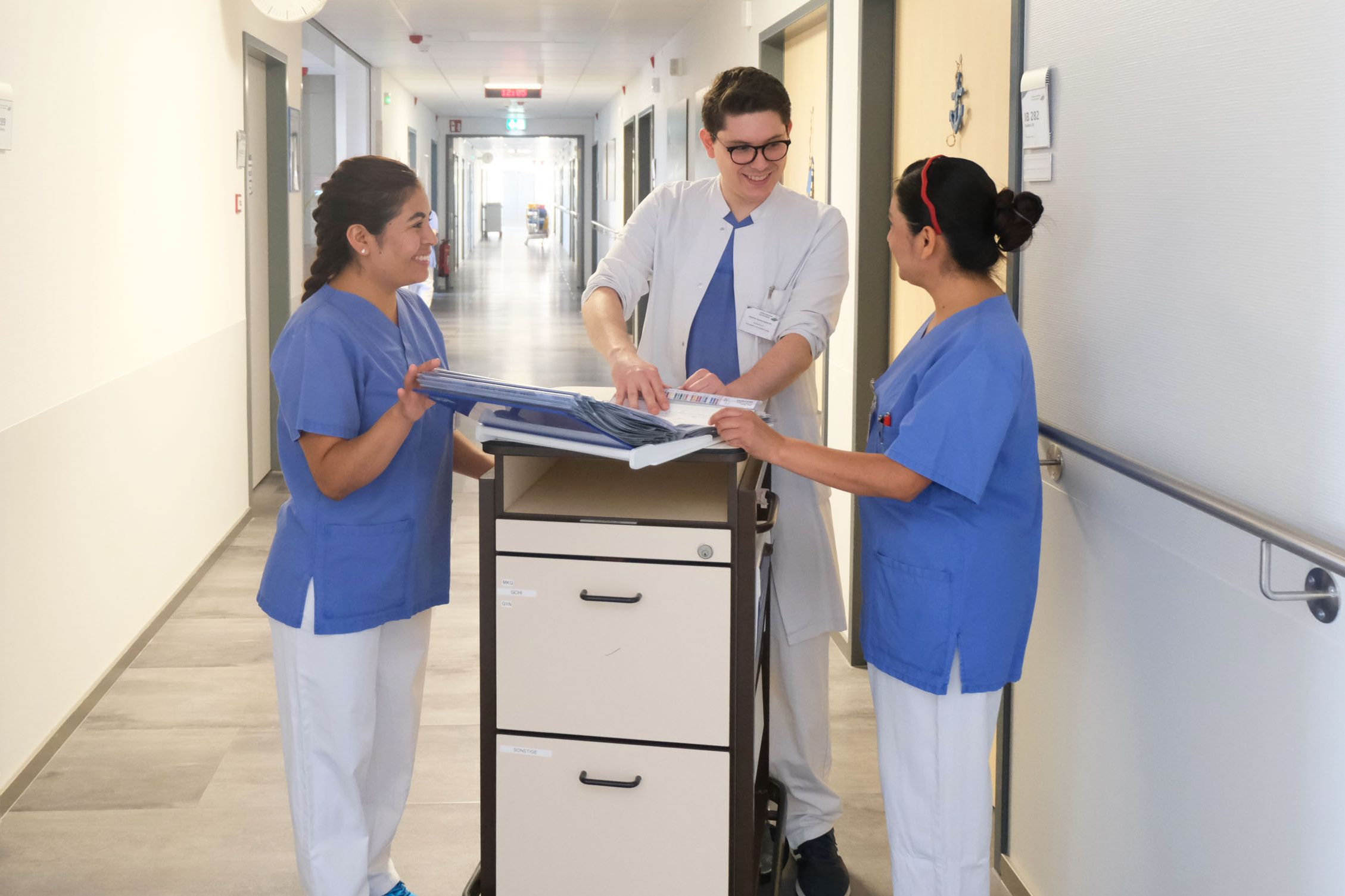 Internationale Fachkräfte im Klinikum Saarbrücken: Visitenvorbereitung - Arzt mit zwei Pflegekräften am Visitenwagen auf dem Flur