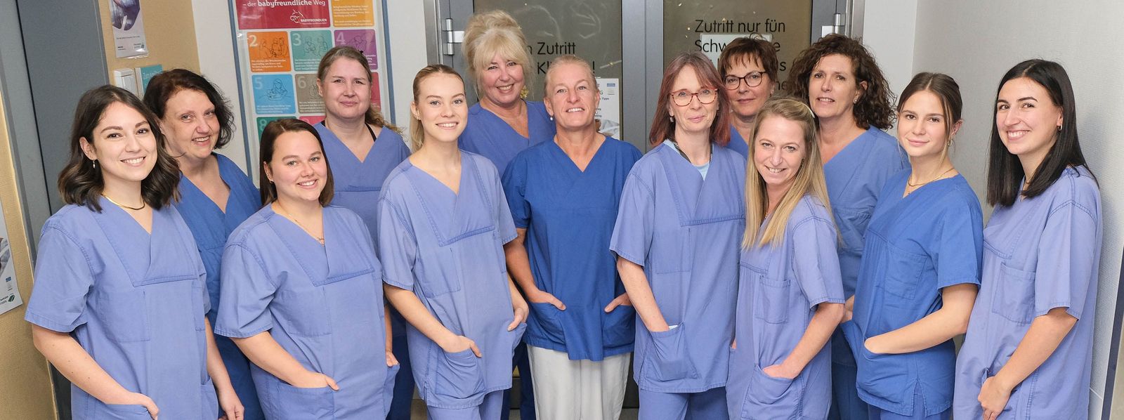 Ein Gruppenfoto unserer Hebammen, sie stehen alle gemeinsam vor der Tür des Kreißsaals Geburtshilfe_Kreisssaal_Teamfoto_Slider.jpg