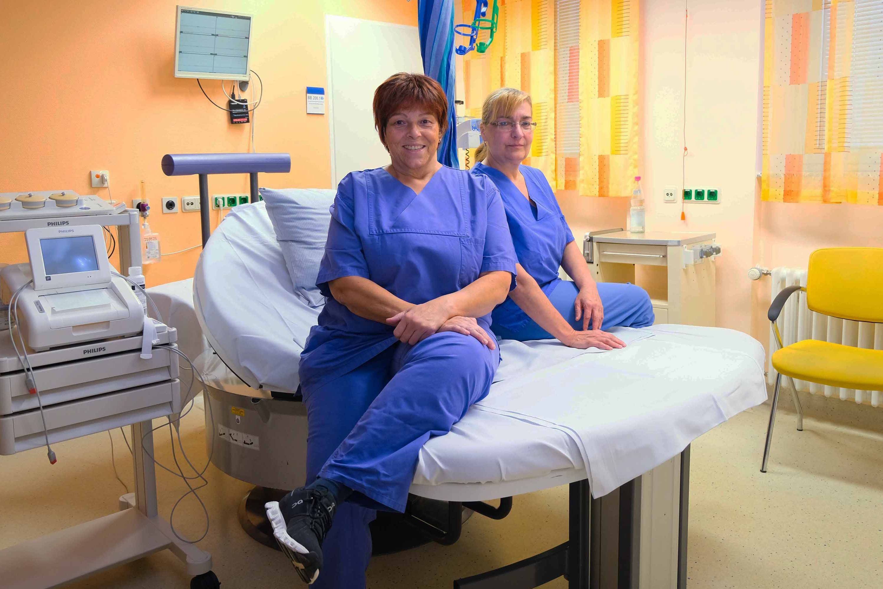 Die beiden Beleghebammen sitzen auf einem Bett im Kreißsaal und lächeln in die Kamera