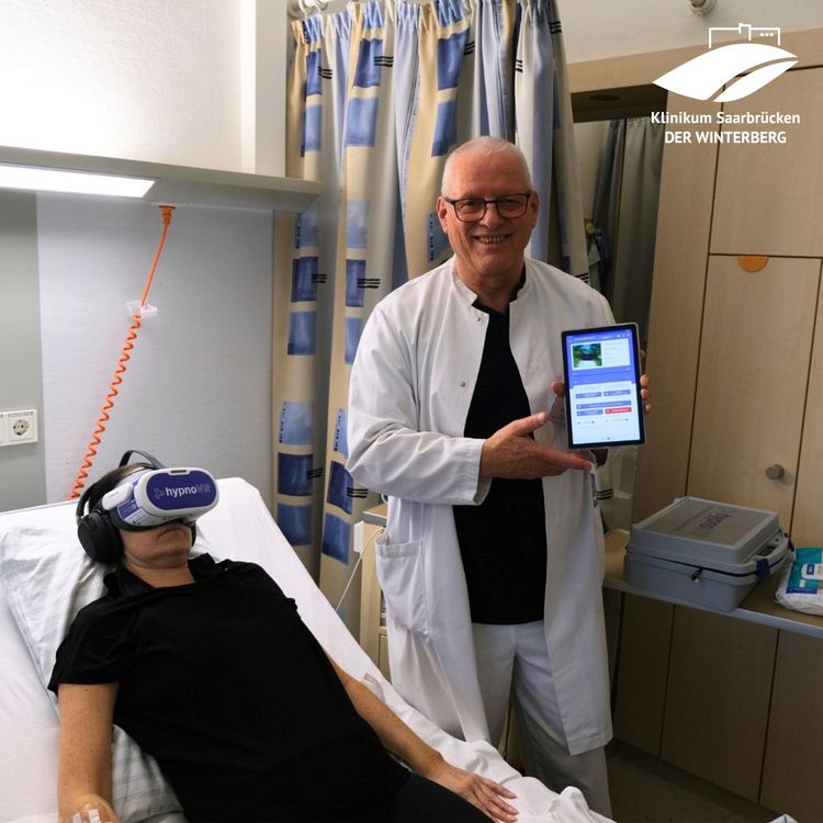 Auf dem Foto sieht man eine Patientin, die im Bett liegt und eine VR-Brille auf dem Kopf trägt. Daneben steht Psychoonkologe Wolfgang Merda und hält ein Tablet in der Hand, mit dem er die VR-Brille steuern kann.