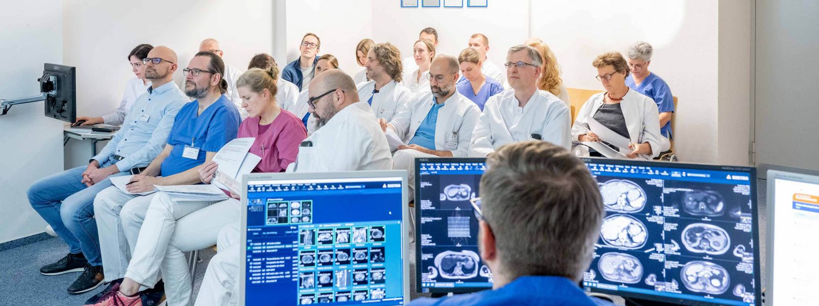 Ein Foto aus der Tumorkonferenz. Darauf zu sehen ist das Team der Allgemeinchirurgie im Hintergrund und im Vordergrund sieht man Bildschirme mit Röntgenbildern Slider_ACHI_3.jpg