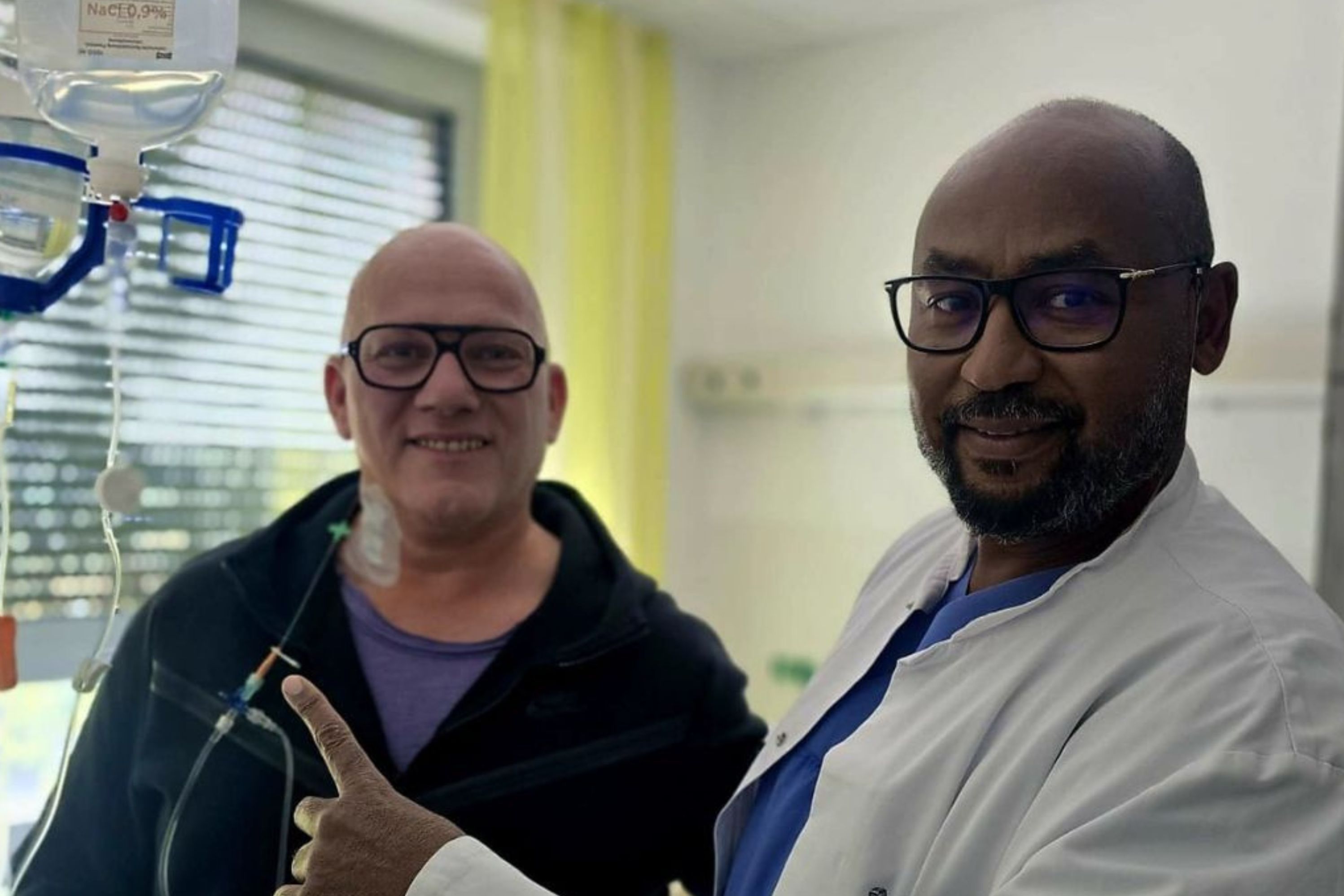 Patient David Saar und Arzt Nadir Tag Elsir schauen in die Kamera und zeigen auf den Chemo-Tropf.