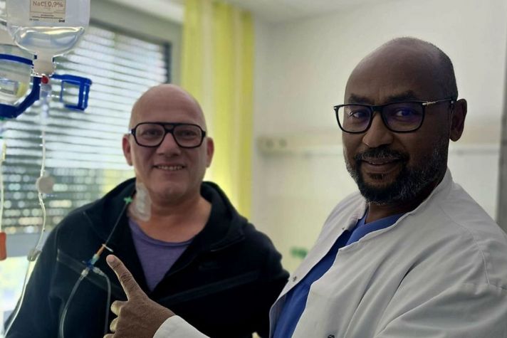 Patient David Saar und Arzt Nadir Tag Elsir schauen in die Kamera und zeigen auf den Chemo-Tropf.