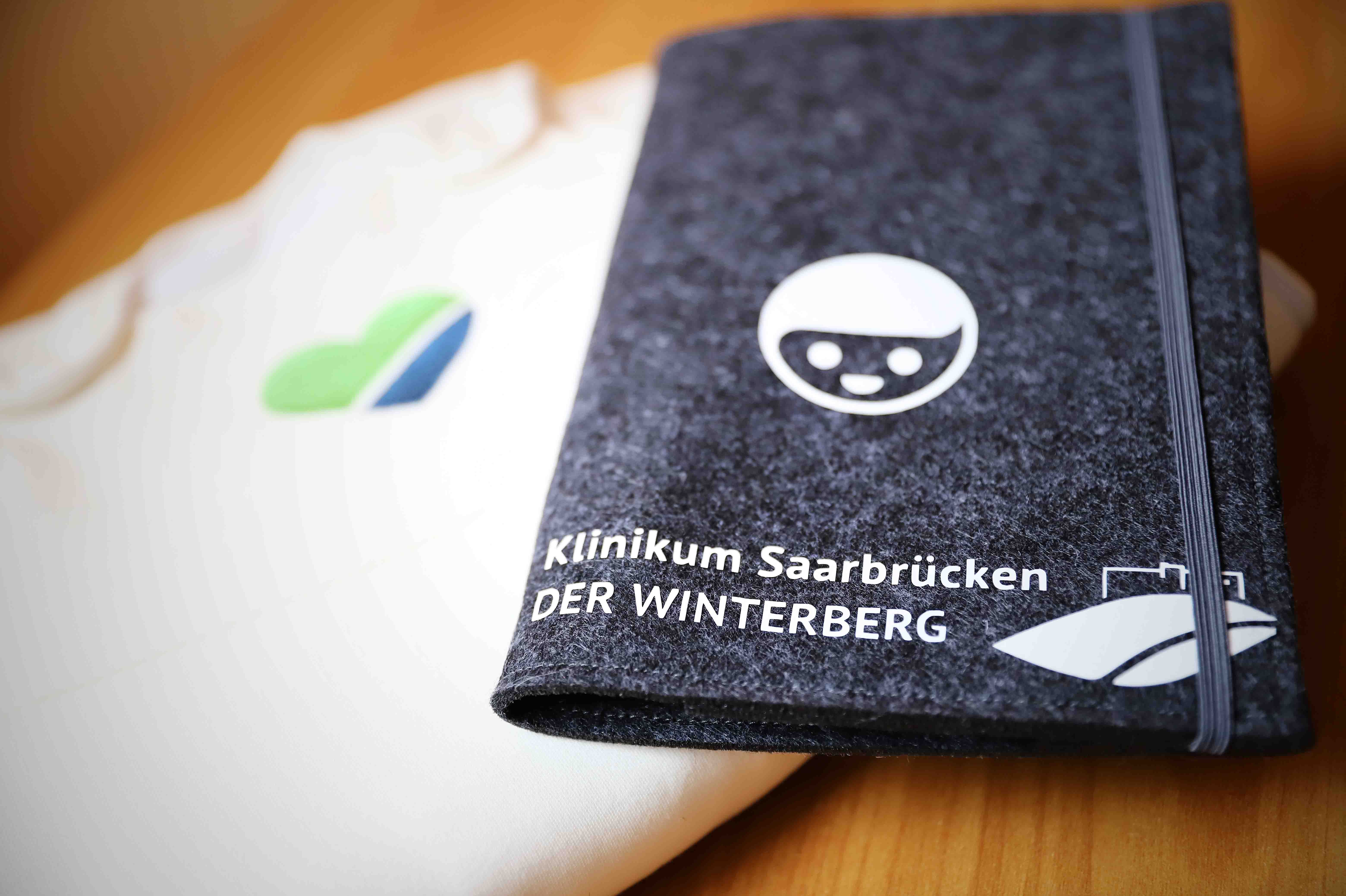 Auf einem Tisch liegen ein Strampler mit einem Herz in den Winterbergfarben sowie die Hülle eines Geburtspasses mit dem Logo des Klinikums Saarbrücken