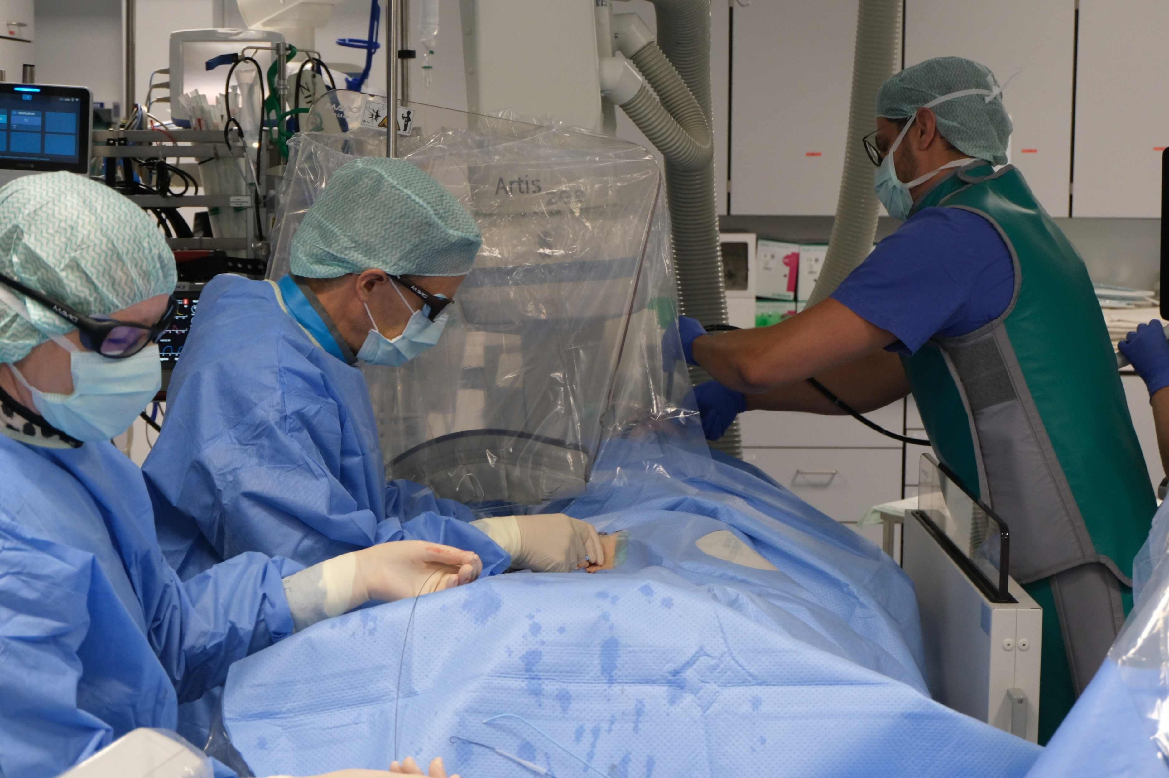 Mehrere medizinische Fachkräfte führen in einem Herzkatheterlabor einen minimalinvasiven Eingriff durch. Die Patientin oder der Patient ist steril abgedeckt. Das Behandlungsteam arbeitet an einem Katheter unter Röntgenbildgebung; im Hintergrund sind medizinische Geräte und Monitore sichtbar.