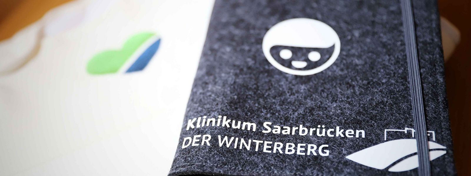 Auf dem Foto sieht man einen Babystrampler mit einem grün blauen Herz und eine grau Mappe auf der Klinikum Saarbrücken steht Slider_Elternschule.jpg