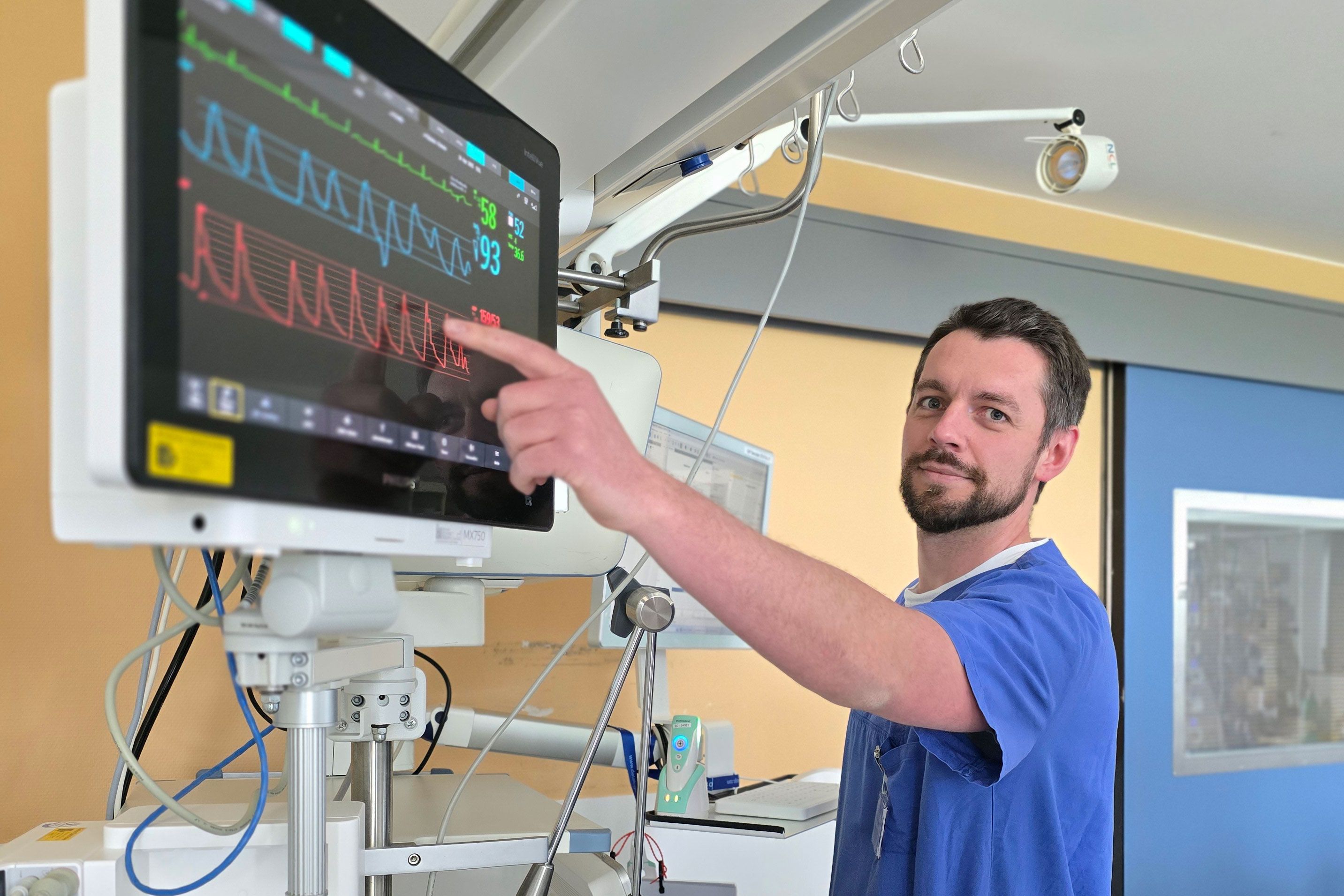 Stationsleitung Karsten Schmitt zeigt auf einen Überwachungsmonitor am Patientenbett.
