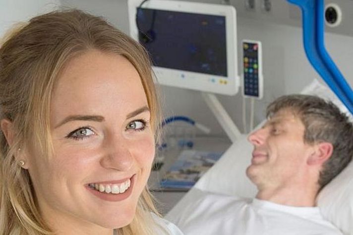 Pflegefachkraft laechelt in die Kamera, Patient im Hintergrund