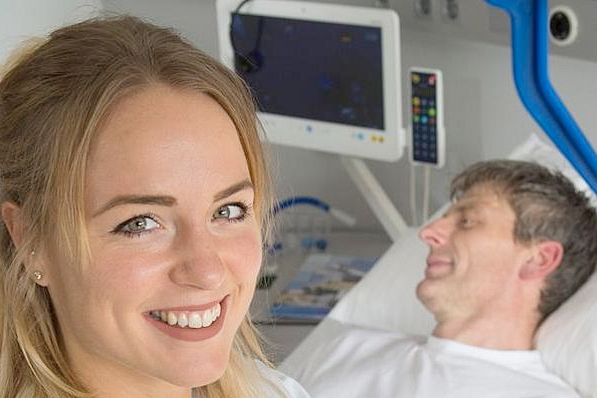 Pflegefachkraft laechelt in die Kamera, Patient im Hintergrund