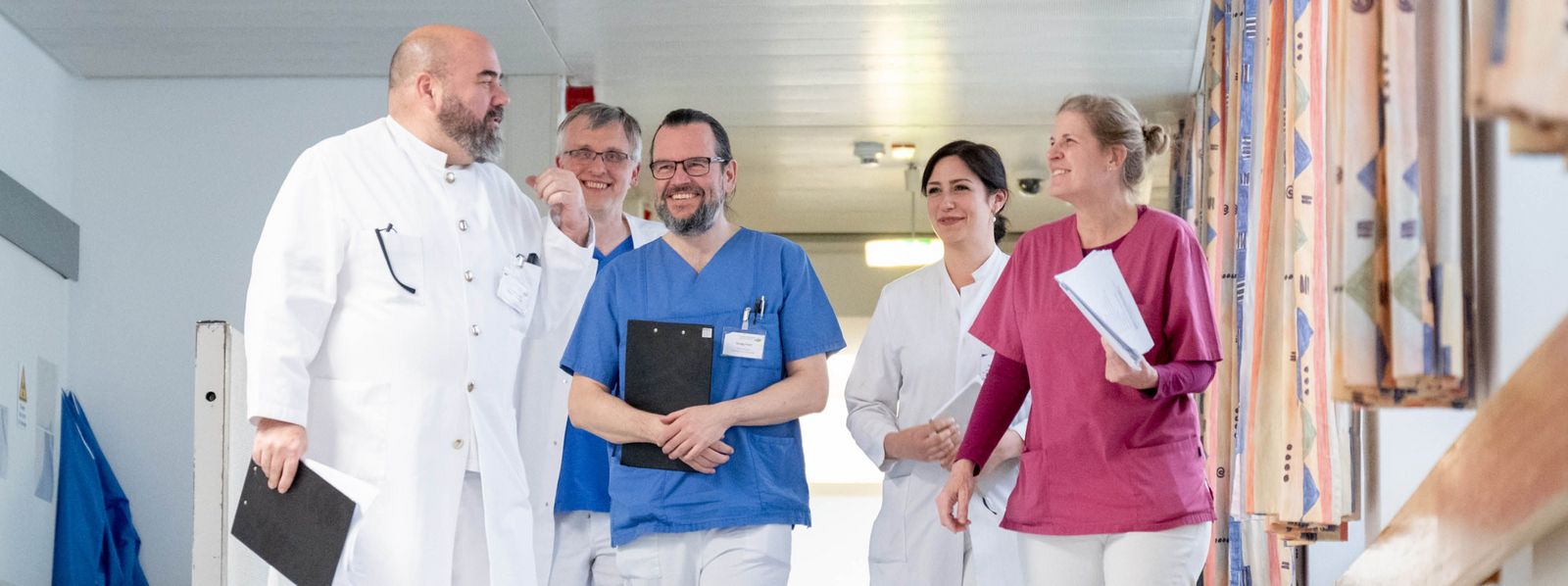Eine Gruppe von Ärzten sowie medizinischem Fachpersonal gemeinsam im Flur bei einer Besprechung Interdisziplinaeres_Arbeiten_Slider.jpg
