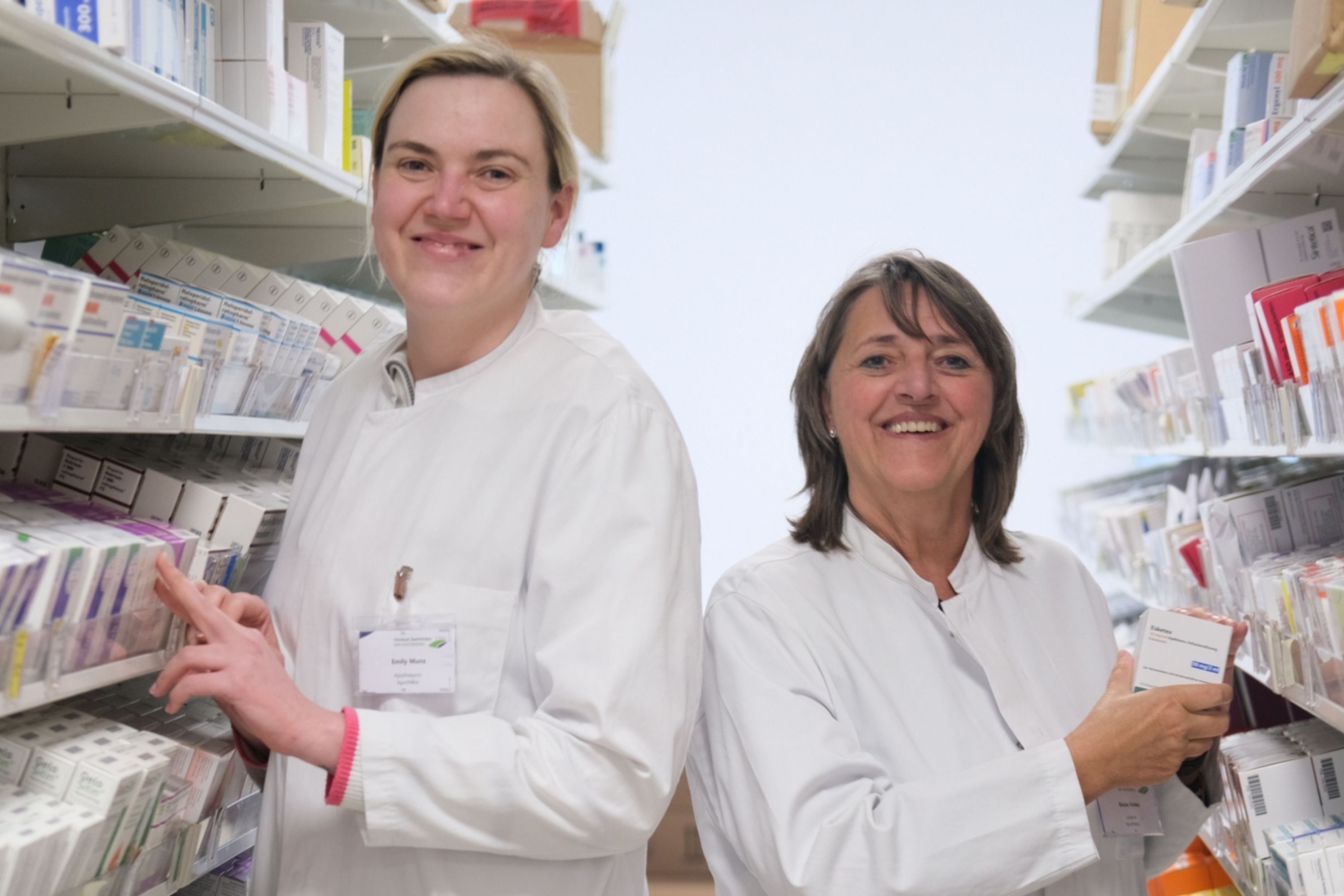 Emily Munz (links) und Beate Halex leiten die Krankenhausapotheke auf dem Winterberg. Sie sind zu sehen, wie sie inmitten von Medikamentenregalen stehen.