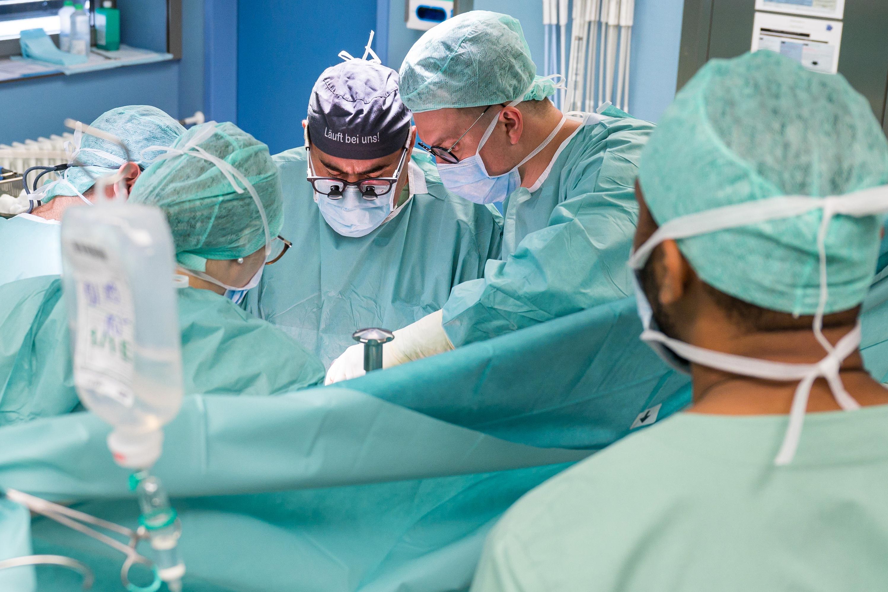 Dr. Dr. Gregor Stavrou mit der OP-Haube "Läuft bei uns" im Operationssaal mit anderen Operateuren. 