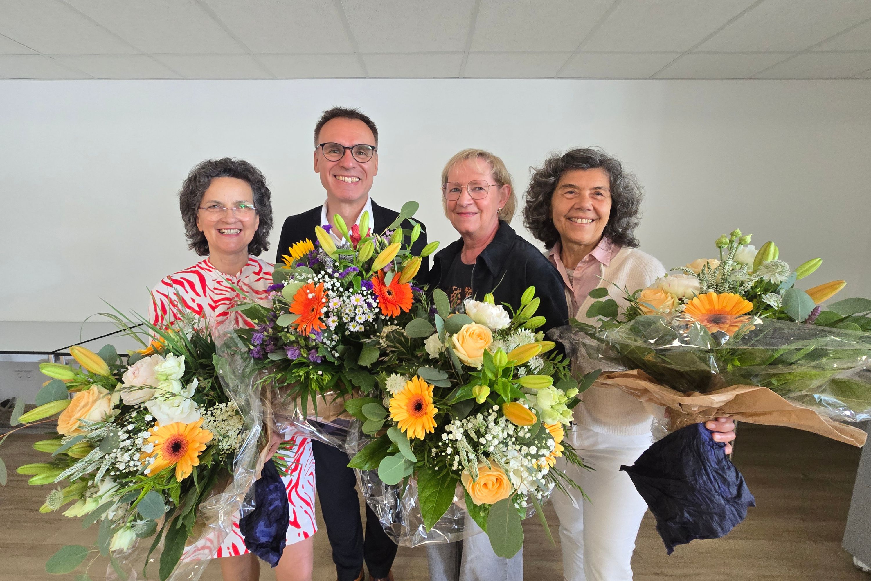 Drei Frauen und ein Mann stehen mit Blumensträußen nebeneinander und lachen in die Kamera.